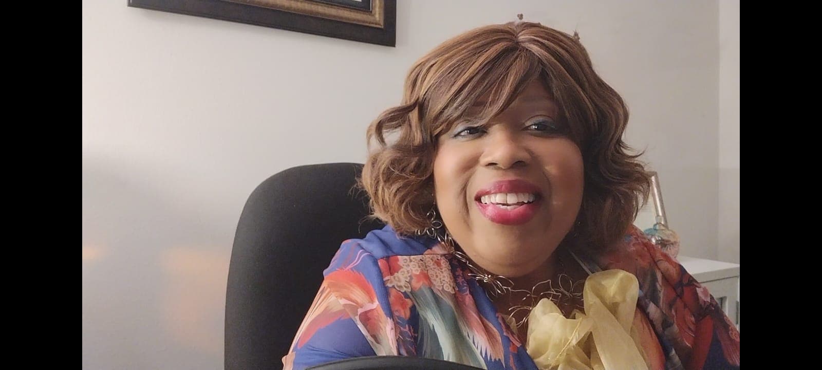 Smiling Black woman with wavy brown hair wearing a colorful floral blouse and yellow ruffles.