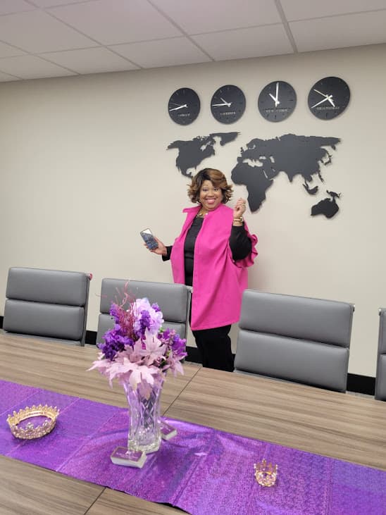 Smiling woman in pink jacket stands in conference room with world map and purple flowers.